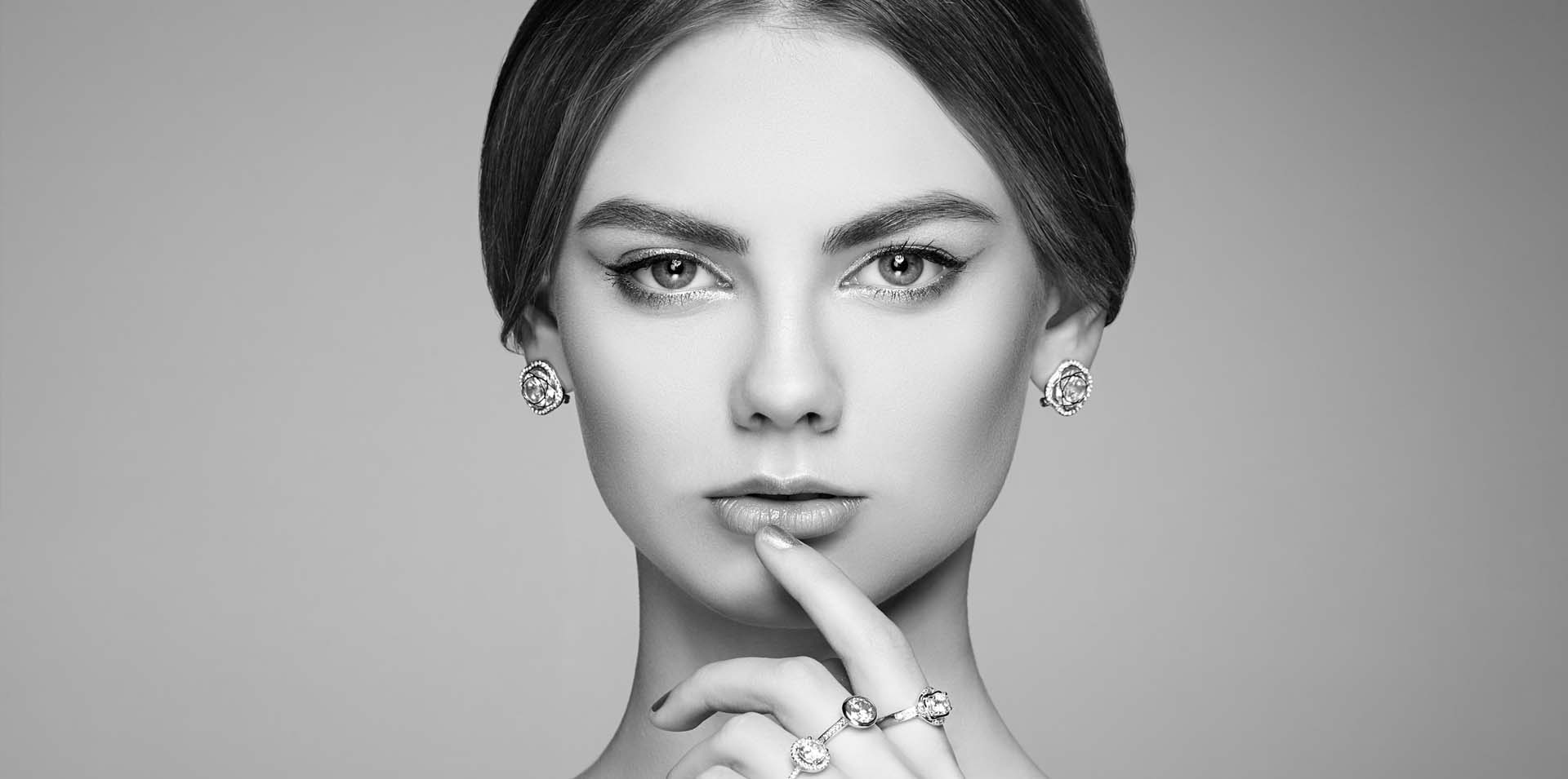 Close up of a woman holding her finger to her mouth. The woman is wearing a pear of diamond earrings and a set of diamond rings on her hands: Telephone number 1-877-872-9909
